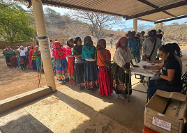 Un grupo de personas vestidas con ropa tradicional hacen fila al aire libre, frente a una mesa de registro, donde dos trabajadores sentados los ayudan.