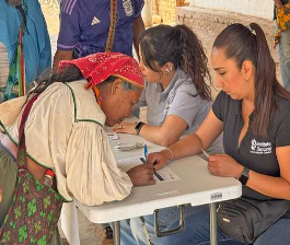Una mujer mayor firma papeles en una mesa mientras dos miembros del personal la ayudan y hay otras personas cerca.