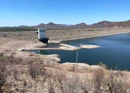 Un embalse con niveles bajos de agua, que muestra una costa expuesta, vegetación seca en primer plano y una estructura de presa bajo un cielo azul claro.