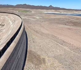 Una presa de hormigÃ³n bordea un embalse prÃ¡cticamente seco, con suelo expuesto y agua lejana bajo un cielo despejado.