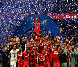 Un equipo de fútbol con uniformes rojos celebra una victoria en el campeonato, sosteniendo un trofeo en medio de confeti que cae sobre un escenario decorado.