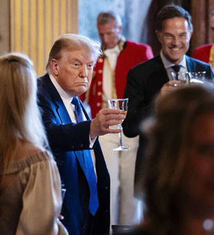 Un hombre con traje azul levanta una copa para brindar en un acto formal en un interior, con otras personas a su alrededor que también sostienen copas.