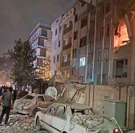 Autos dañados y escombros se alinean en la calle afuera de un edificio de varios pisos con fuego y destrucción visibles en algunos departamentos por la noche.