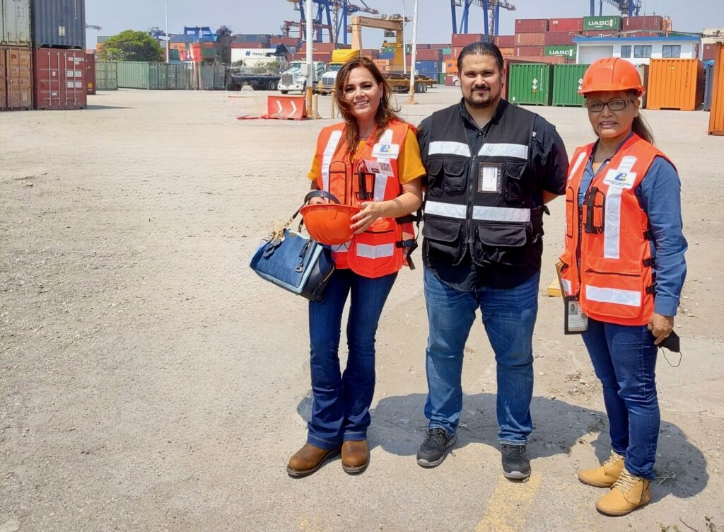 Tres personas que visten chalecos de seguridad y cascos se encuentran paradas en un lote de grava en un patio de contenedores de envío, con contenedores apilados y grúas en el fondo.