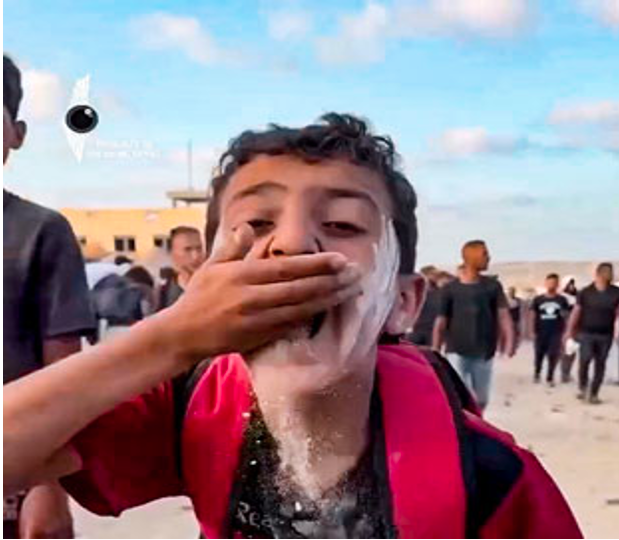 Un niño con harina o polvo en la cara se tapa la boca con la mano mientras está al aire libre entre un grupo de personas.