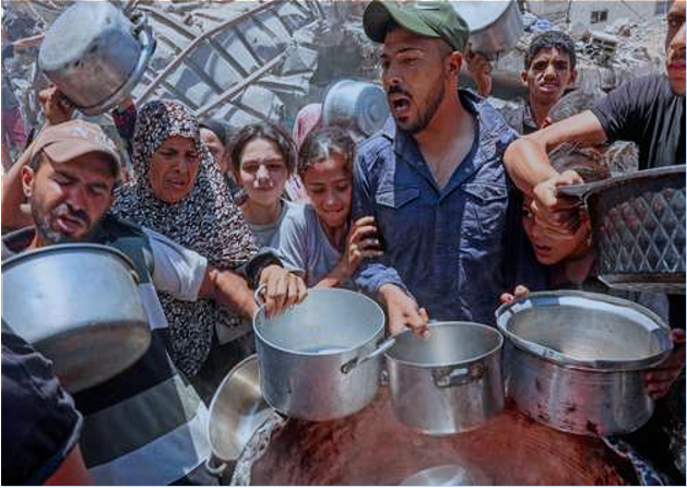 Un grupo de personas se agolpa con ollas metálicas, alcanzando alimentos que se distribuyen al aire libre en un entorno abarrotado y caótico.