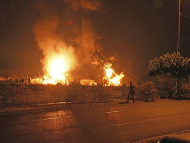 Grandes llamas y humo se elevan desde estructuras en llamas por la noche, con siluetas de personas visibles cerca del incendio y una carretera en primer plano.