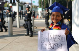 Una joven con toga y birrete de graduación de color azul sostiene un cartel que dice "Por mamá, por papá", mientras al fondo hay agentes de policía.