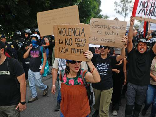 Manifestantes con pancartas en español contra la violencia y el aburguesamiento, con agentes de policía visibles al fondo.