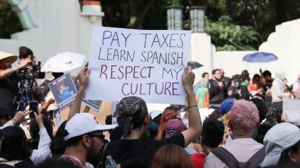 Una persona entre la multitud sostiene un cartel en el que se lee: "Paga impuestos, aprende español, respeta mi cultura", durante una manifestación pública.