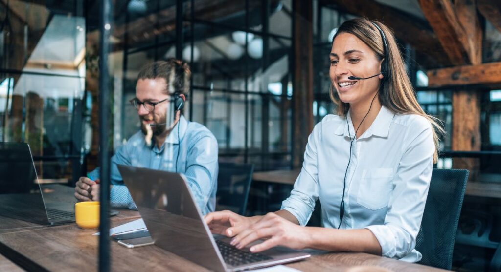 Dos personas con auriculares trabajan con ordenadores portÃ¡tiles en una oficina moderna con paredes de cristal y vigas de madera a la vista, colaborando para atender a el cliente con eficacia.