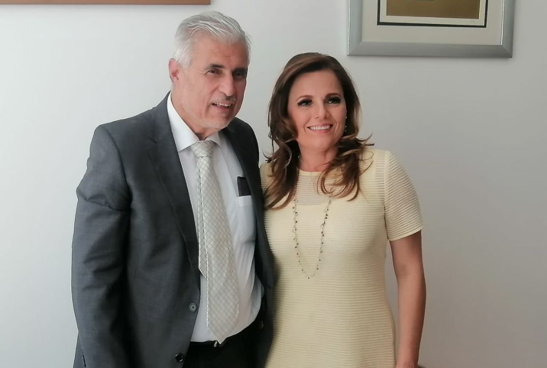 Dos adultos en un interior, uno con traje gris y corbata y el otro con un vestido claro. Ambos sonríen y miran a la cámara.