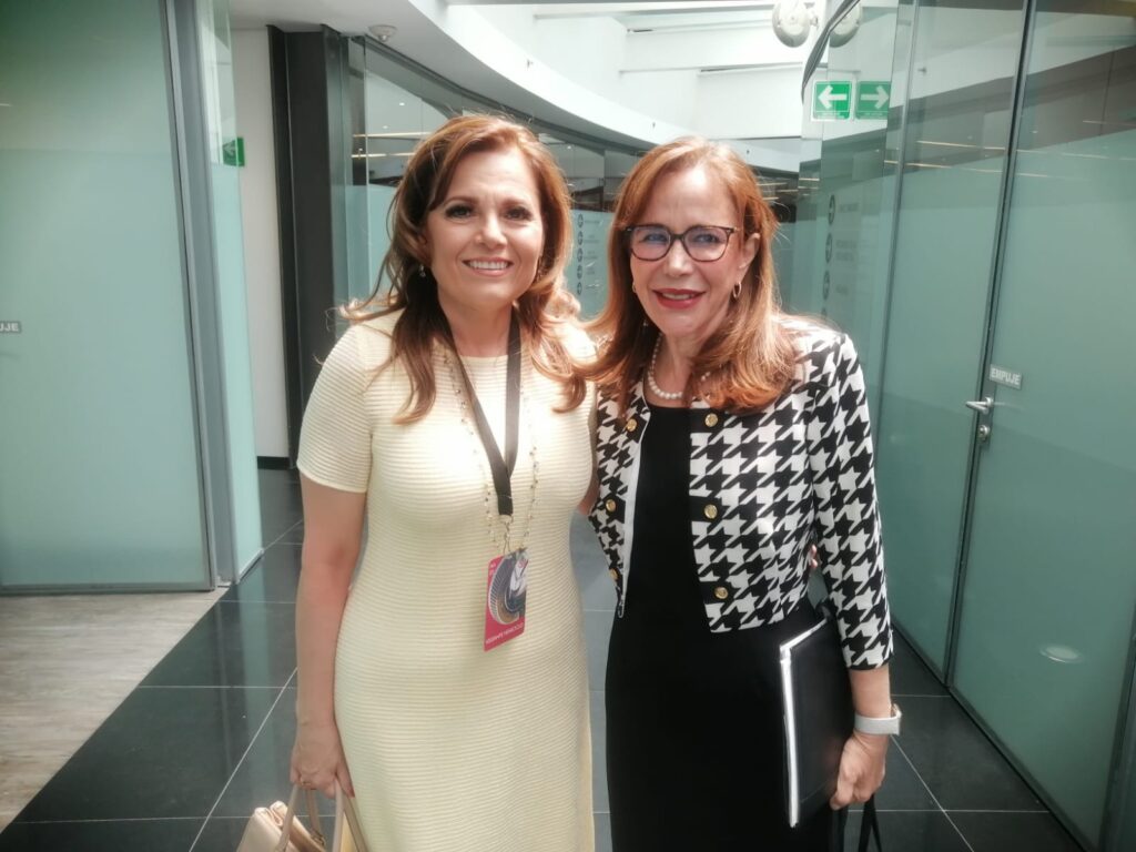 Dos mujeres de pie en el interior, sonriendo a la cámara; una lleva un vestido amarillo claro y la otra una chaqueta de pata de gallo en blanco y negro, con una carpeta en la mano.