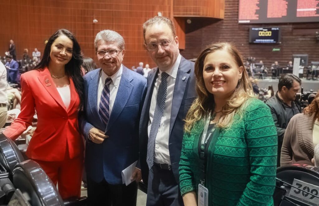 Cuatro personas posan juntas y sonríen para una foto en un amplio espacio interior con paredes de paneles de madera y pantallas electrónicas de fondo.