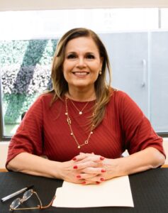 Una mujer de pelo castaño con top rojo y collar dorado se sienta en una mesa con las manos cruzadas, una carpeta y gafas, sonriendo a la cámara.