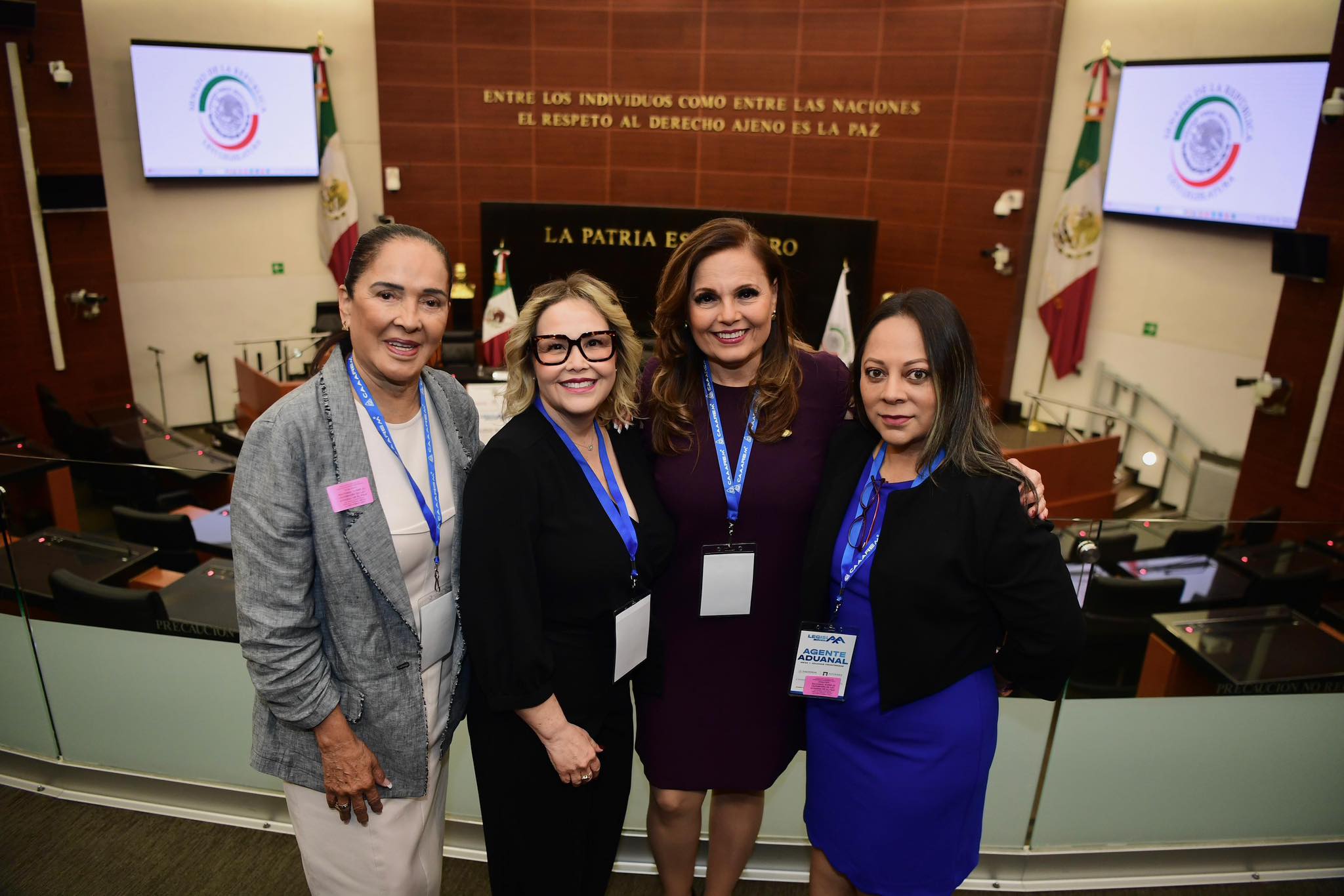 Cuatro mujeres, entre ellas Laura Blanco, con insignias de la conferencia, se paran juntas y sonríen para una foto en una sala de reuniones formal con banderas y emblemas oficiales, destacando su experiencia compartida en importación y exportación.