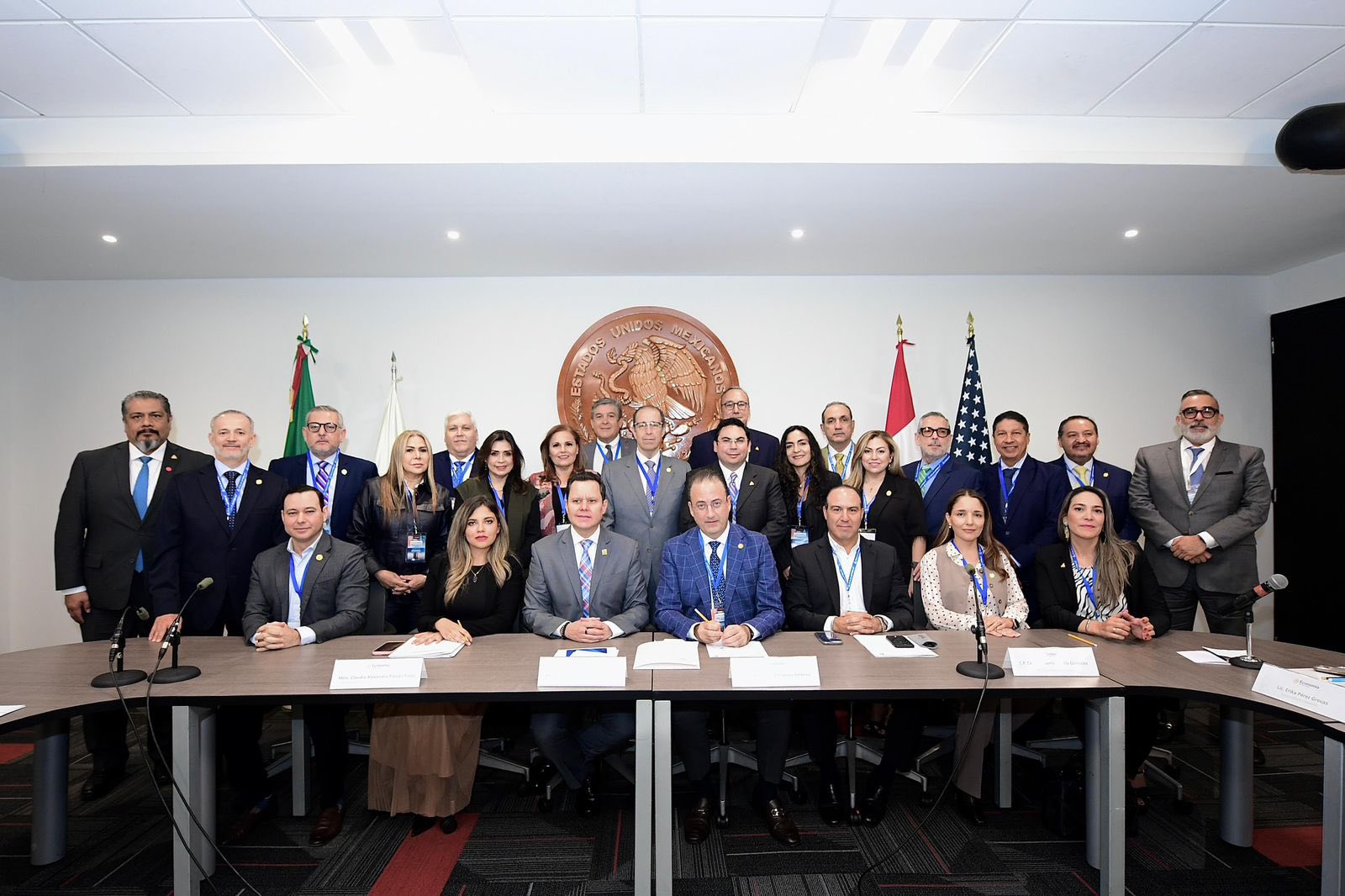 Un grupo de personas en traje de etiqueta posa para una foto detrás de una mesa de conferencias con documentos, banderas y un gran sello oficial en la pared, en la que destacan los debates sobre importación y almacenaje dirigidos por Laura Blanco.