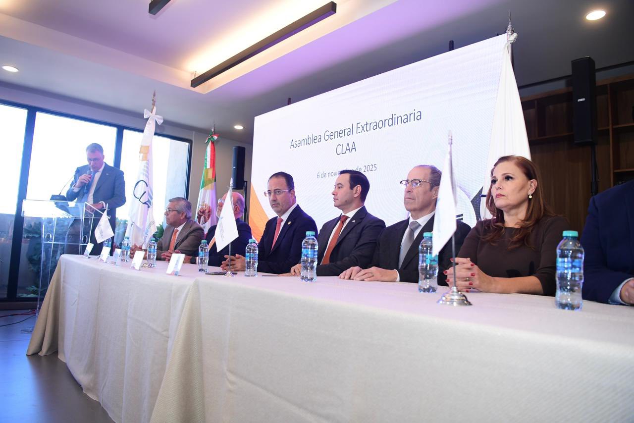 Un grupo de personas vestidas de etiqueta están sentadas en una larga mesa con micrófonos y botellas de agua, mientras un hombre habla en un podio al fondo durante una asamblea oficial centrada en la importación y la exportación.