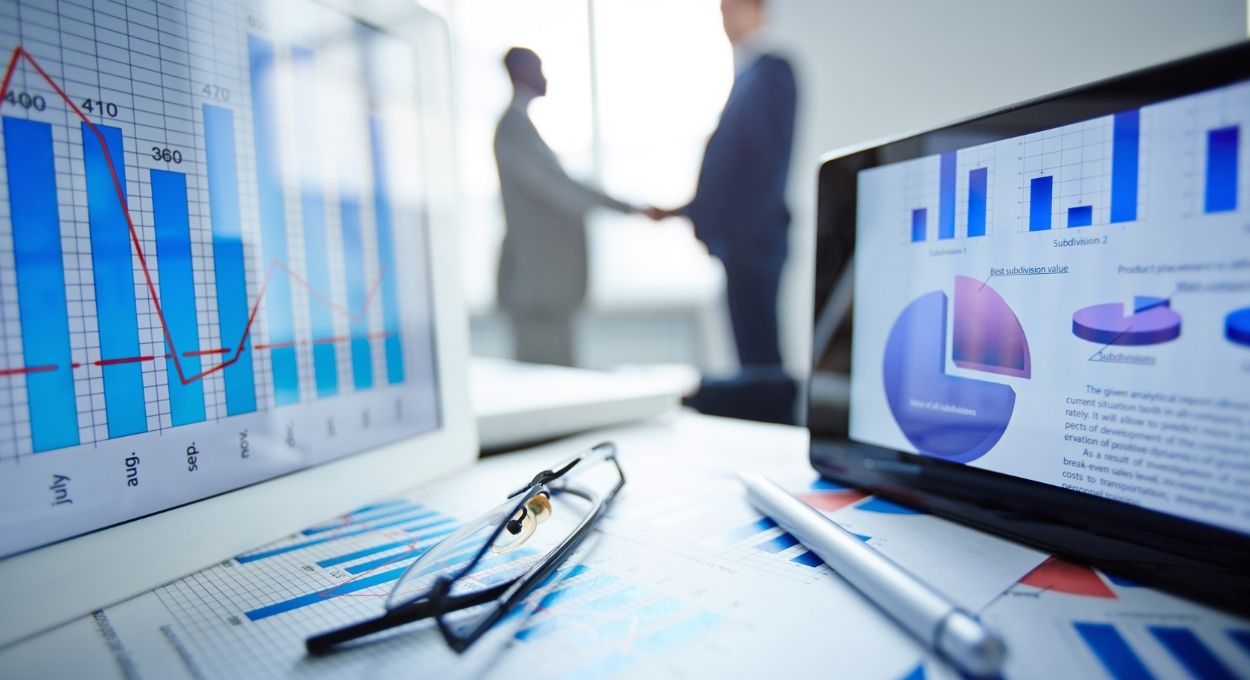 Dos personas se dan la mano en una oficina. En primer plano se ven tablas y gráficos relacionados con el mercado interior en papel y pantallas digitales, junto con gafas y un bolígrafo sobre un escritorio.