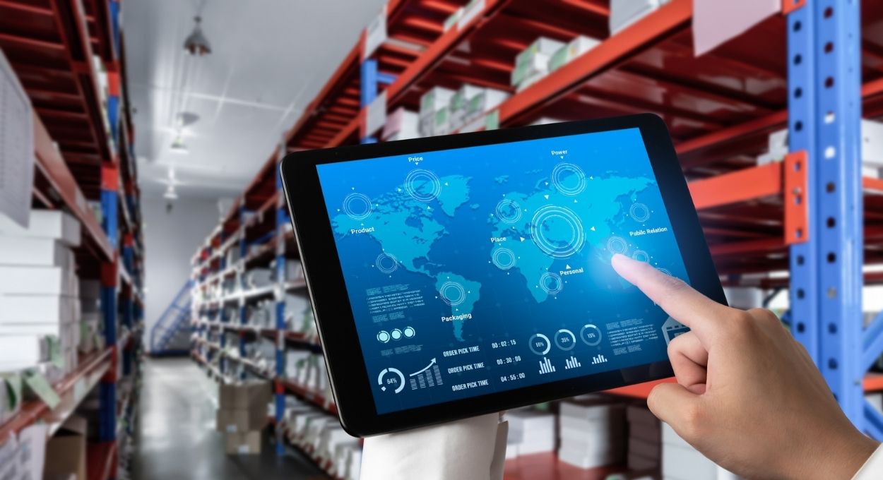 Una persona utiliza una tableta que muestra un mapamundi y datos logísticos en el pasillo de un almacén lleno de estanterías y cajas, optimizando las importaciones inteligentes.