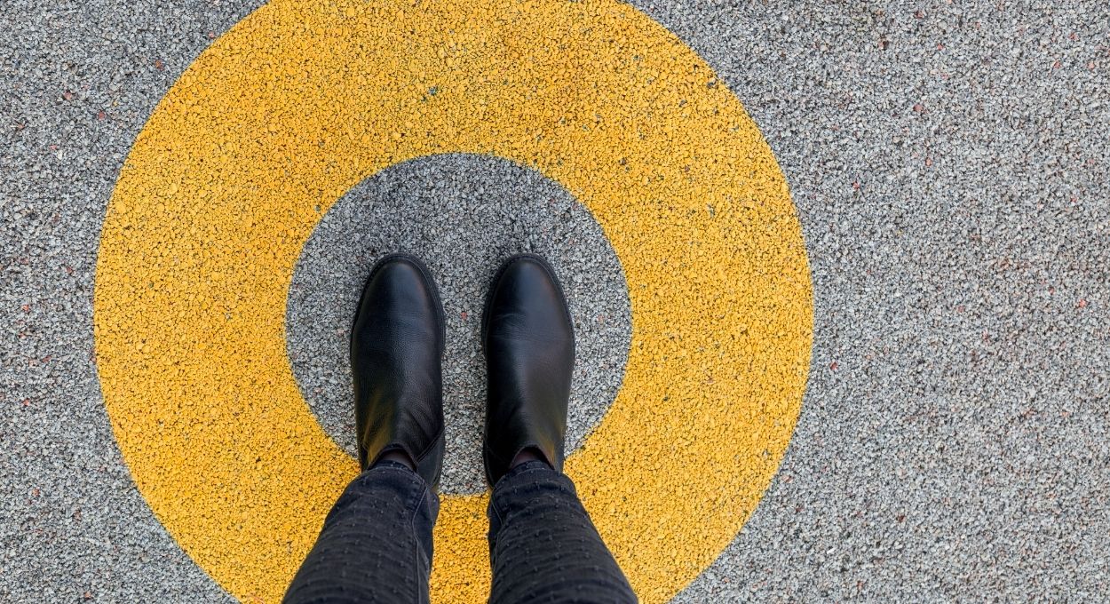 Una persona con botas negras de pie en el centro de un círculo amarillo pintado sobre una superficie gris con textura.